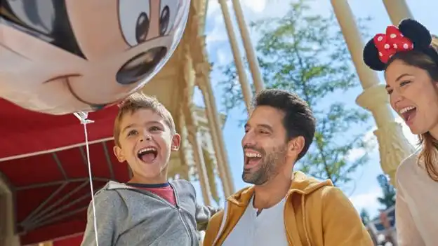 Famille heureuse à Disneyland Paris, avec un enfant tenant un ballon Mickey et des parents souriants portant des accessoires Disney, profitant d'une journée ensoleillée dans le parc.